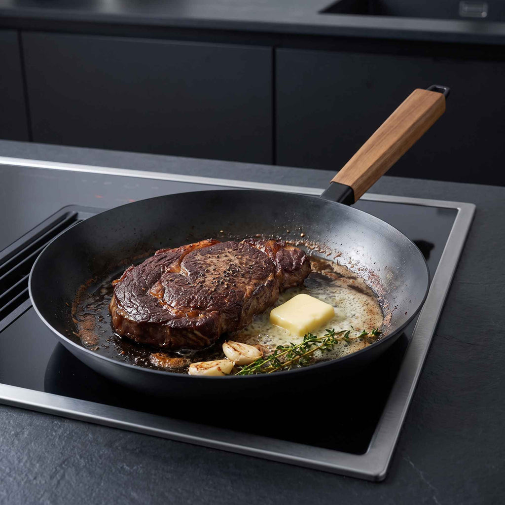 Set of four Alva Cookware carbon steel frying pans, with one searing steak on an induction stove.