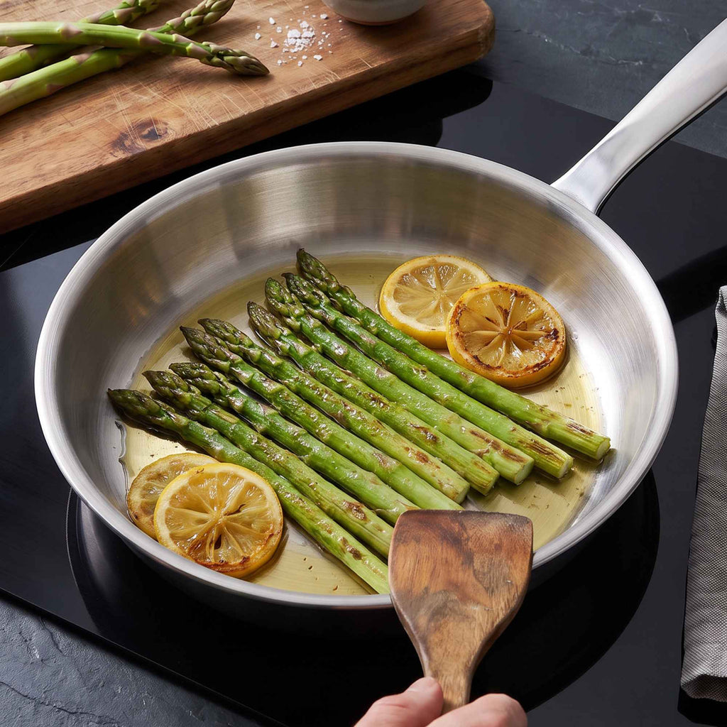 Alva Cookware 5-Ply stainless steel frying pan sautéing asparagus and lemon on a stovetop.